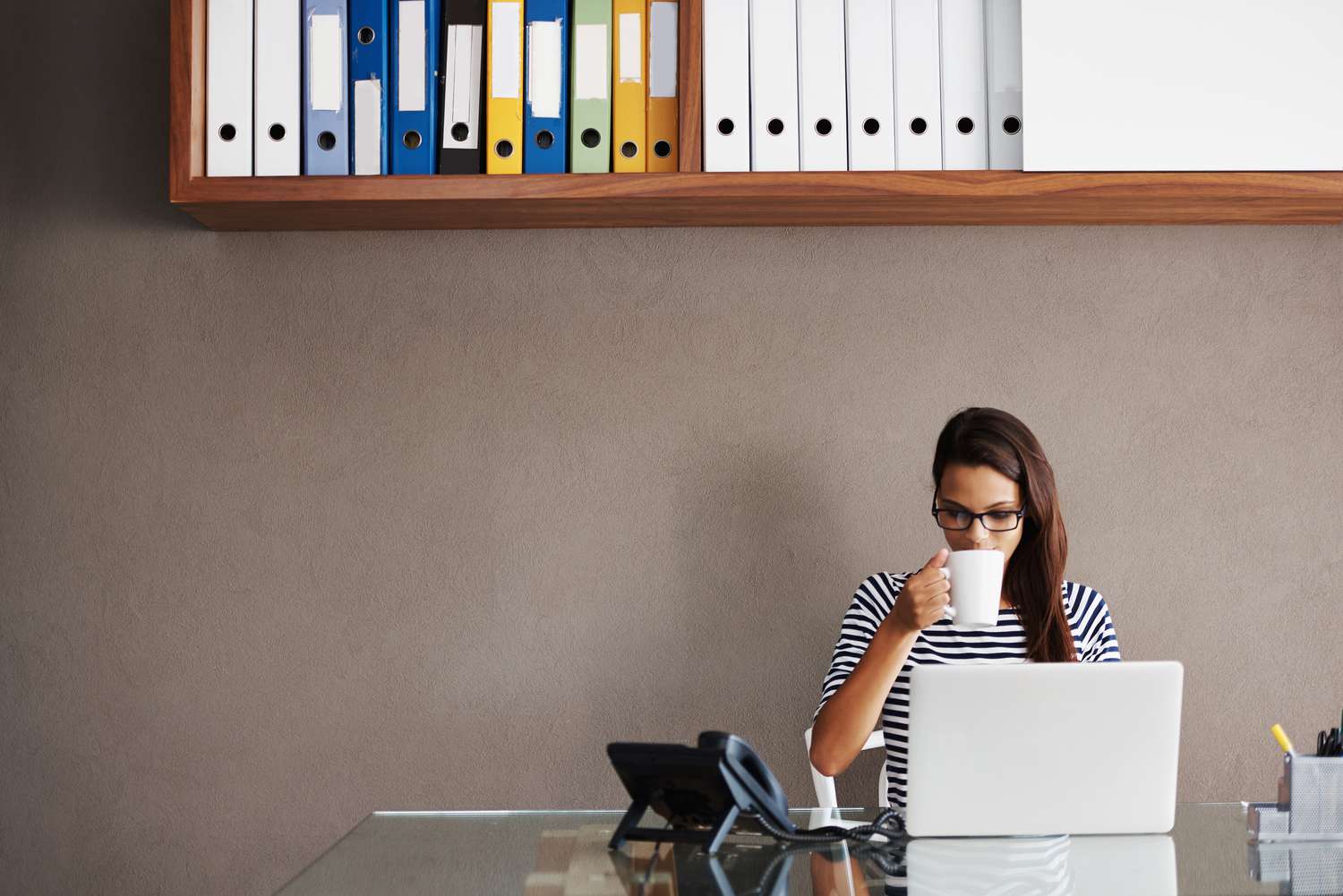 Personal assistant sitting at desk drinking coffee and looking at a laptop