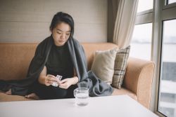 Female sitting on sofa with sickness cover with blanket taking medicine