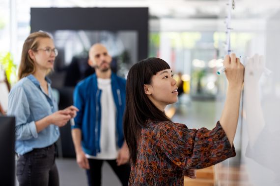 Young businesswoman writes on whiteboard as two colleagues watch