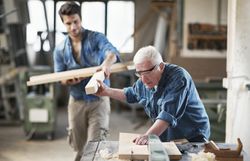 Construction helper handing a piece of wood to a carpenter working at a table saw in a shop.