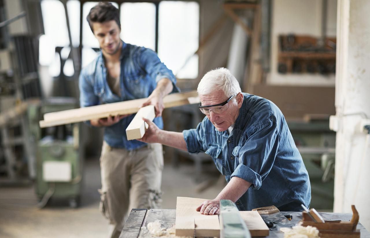 Construction helper handing a piece of wood to a carpenter working at a table saw in a shop.