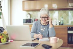 Older woman on phone writes on notepad in kitchen