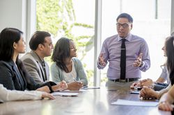 Man standing at conference table gives presentation to colleagues