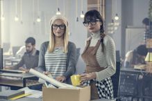 Two young women, a blond and a brunette, are packing up their office coffee cup and papers into a brown box because they were laid off.