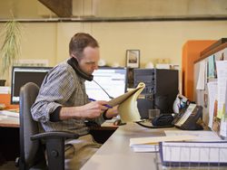 Manager at his desk reviewing reports while talking on the phone at the same time.